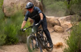 Una persona que lleva casco y gafas de sol va en bicicleta de montaña por un sendero de tierra rodeado de rocas y vegetación.