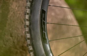 Primer plano de una rueda y una llanta de bicicleta de montaña en las que se aprecia el logotipo «ESC», situadas junto a una gran roca en un entorno al aire libre.