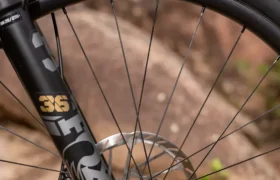 Primer plano de la rueda delantera de una bicicleta de montaña con un neumático Maxxis, un freno de disco y una horquilla de suspensión FOX 36, con un fondo rocoso al aire libre.