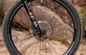 Un primer plano de la rueda delantera de una bicicleta de montaña con un neumático Maxxis, un freno de disco y una horquilla de suspensión, situada en un sendero de tierra con rocas y vegetación al fondo.