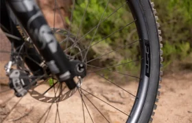 Primer plano de la rueda delantera de una bicicleta con freno de disco y el logotipo de FSA, aparcada en un camino de tierra con vegetación al fondo.