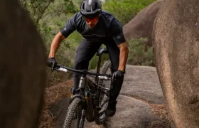 Un ciclista de montaña, con casco y gafas de sol, pedalea sobre las rocas por un sendero de tierra rodeado de grandes cantos rodados y vegetación.