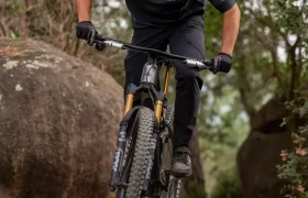 Una persona que lleva casco y gafas de sol va en bicicleta de montaña por encima de una gran roca, rodeada de árboles y cantos rodados.
