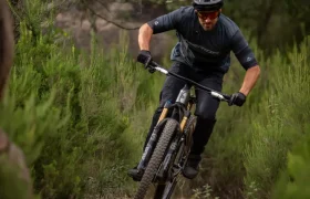 Una persona que lleva casco y gafas de sol baja en bicicleta de montaña por un sendero de tierra rodeado de arbustos verdes y árboles.