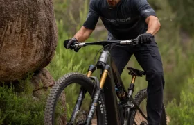 Una persona que lleva casco y gafas de sol va en bicicleta de montaña por un sendero rocoso rodeado de vegetación y grandes rocas.
