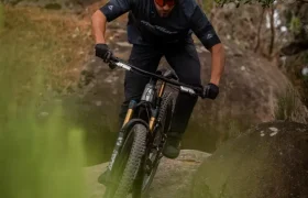 Un ciclista de montaña, vestido con ropa oscura y casco, baja por una pendiente rocosa por un sendero forestal, rodeado de vegetación y árboles.