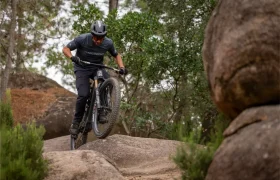 Una persona que lleva casco y gafas de sol va en bicicleta de montaña por unas rocas grandes en un sendero rodeado de árboles y arbustos.