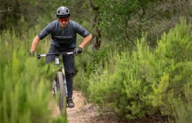 Una persona que lleva casco y gafas de sol va en bicicleta de montaña por un sendero de tierra rodeado de arbustos y árboles verdes.