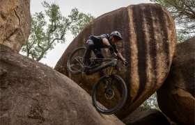 Una persona que lleva casco baja en bicicleta de montaña por una pendiente empinada y rocosa, con grandes rocas y árboles al fondo.