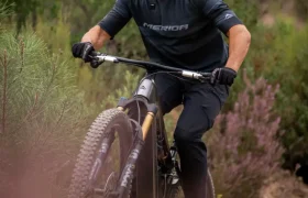 Una persona que lleva casco y gafas de sol sube en bicicleta de montaña por un sendero de tierra rodeado de vegetación y pequeños arbustos.