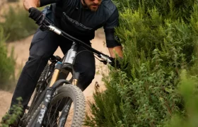 Una persona que lleva casco y gafas de sol va en bicicleta de montaña por un sendero de tierra, inclinándose en una curva cerca de unos arbustos verdes.
