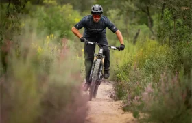 Una persona que lleva casco monta en bicicleta de montaña por un sendero de tierra rodeado de vegetación y flores silvestres.