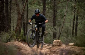 Una persona que lleva casco y gafas de sol sube en bicicleta de montaña por un sendero rocoso en una zona boscosa.