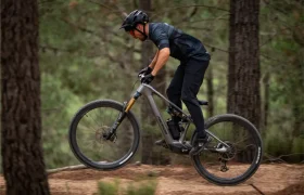 Una persona que lleva casco y ropa oscura circula en bicicleta de montaña por un sendero de tierra en una zona boscosa, levantando la rueda delantera del suelo.