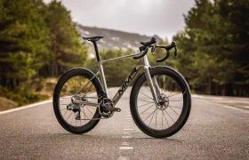Una bicicleta de carretera MMR Aelion plateada con ruedas negras y manillar caído está aparcada en una carretera asfaltada vacía bordeada de árboles y montañas al fondo.