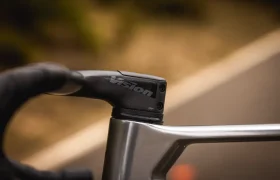 Una mano enguantada agarra la potencia de un manillar Vision en una MMR Aelion SL, sujeta a un cuadro de bicicleta metálico, con una carretera borrosa de fondo: perfecto para cualquier entusiasta de la bici total.