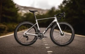 Una bicicleta de carretera MMR Aelion plateada con manillar rebajado y ruedas de sección profunda está situada en la línea central de una carretera vacía, rodeada de árboles.