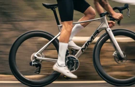 Una persona vestida de ciclista conduce a toda velocidad una bicicleta de carretera MMR Aelion SL plateada por una carretera asfaltada, con desenfoque de movimiento de fondo, mostrando las prestaciones de esta bici asturiana.