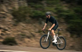 Una persona con casco y ropa de ciclismo monta en una bicicleta de carretera MMR Aelion SL por una carretera asfaltada con un fondo natural difuminado, mostrando las prestaciones del equipamiento bici total.