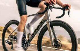 Persona montando en una bicicleta de carretera MMR Aelion con la marca MMR en un camino pavimentado al aire libre, luciendo ropa de ciclismo que incluye culotte, maillot y calcetines blancos, perfecta para sentir la experiencia total de la bicicleta.
