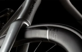 Primer plano de un cuadro de bicicleta MMR Aelion plateado y una rueda de carbono, destacando la marca de la cubierta y parte de la horquilla de la bicicleta, sobre un fondo oscuro.