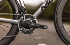 Primer plano de unas bielas y un plato de bicicleta Aelion SL fijados a un cuadro de bicicleta plateado, con parte de la rueda trasera y el neumático visibles.