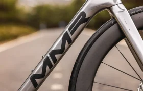 Primer plano de un cuadro plateado de bicicleta MMR Aelion SL con el logotipo "MMR", mostrando la rueda delantera y la horquilla, colocado en una carretera asfaltada con fondo borroso.