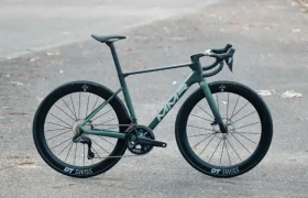 Una bicicleta de carretera MMR Aelion verde mate con ruedas DT Swiss negras está aparcada en una carretera vacía, pavimentada en curva y con hierba a los lados, encarnando ese espíritu bici total y un sutil acento asturiano.