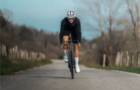 Ciclista con casco y ropa deportiva oscura conduce una bicicleta de carretera MMR Aelion SL por una carretera rural asfaltada con árboles y un cielo nublado de fondo.