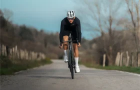 Un ciclista con atuendo negro y casco blanco conduce una bicicleta de carretera MMR Aelion SL por una carretera rural vacía bordeada de árboles y vallas, destacando la velocidad y el estilo de las aventuras bici total.
