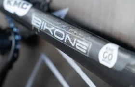 Primer plano de una cadena y un cuadro de bicicleta, mostrando la marca "BIKONE" con una etiqueta "KMC" y una pegatina en la que se lee "EPIC GO original". Las cadenas KMC impulsan al Caja Rural-Seguros RGA en cada trayecto.