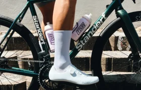Una persona vestida con ropa de ciclista blanca está sentada en una bicicleta Stander verde con dos botellas de agua moradas sujetas al cuadro, situada en unos escalones de piedra.