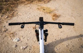Vista desde arriba del manillar de una bicicleta de montaña, que cuenta con un manillar integrado de carbono FSA NS SIC, junto con la potencia y la suspensión delantera, situado en un camino de grava al aire libre, con algo de hierba seca visible al fondo.