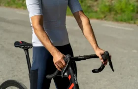 Un ciclista que lleva un casco blanco, gafas de sol y ropa de ciclismo —entre la que destaca el maillot POC Cadence, diseñado para ofrecer una comodidad óptima— aparece de pie en una carretera sosteniendo una bicicleta de carretera negra y roja. El culote Cadence Cargo aporta comodidad al pedalear, garantizando que cada salida sea a la vez elegante y eficiente.
