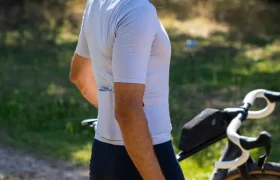 Una persona que lleva un casco polivalente y ropa de ciclismo posa junto a una bicicleta al aire libre en un día soleado, con árboles y césped al fondo, lista para una aventura por caminos de grava o por carretera.
