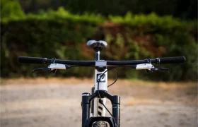 Vista frontal de una bicicleta de montaña equipada con un manillar integrado de carbono FSA NS SIC, de gran anchura, y manetas de freno, aparcada al aire libre sobre tierra, con vegetación al fondo.