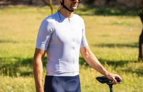 Un ciclista que lleva un casco polivalente blanco, gafas de sol y ropa de ciclismo está al aire libre junto a una bicicleta negra de gravel en un día soleado.