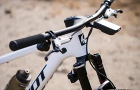 Primer plano del manillar y la suspensión delantera de una bicicleta de montaña Pivot blanca, con un manillar integrado de carbono FSA NS SIC y un portabotellas montado en el cuadro, al aire libre sobre una superficie de tierra.