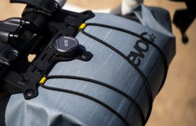 Primer plano de un manillar de bicicleta con una bolsa EVOC Bikepacking de color gris azulado, que ofrece protección y está sujeta por correas negras y un sistema de cierre Boa, sobre un fondo borroso de exteriores.