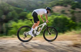 Una persona que lleva casco y equipación de ciclismo recorre a gran velocidad una bicicleta de montaña KFX i30 por un camino de tierra, con árboles verdes al fondo, manteniendo un control total sobre el terreno accidentado gracias a las ruedas FSA de alto rendimiento.