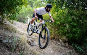 Una persona que lleva casco y gafas de sol desciende en una bicicleta de montaña KFX i30 por un sendero de tierra rodeado de árboles y arbustos, manteniendo con seguridad el control total sobre el terreno accidentado.
