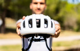 Una persona que sostiene un casco de bicicleta blanco «casco polivalente» hacia la cámara con los brazos extendidos, al aire libre con un fondo verde difuminado: protección perfecta tanto si vas por grava como por carretera.