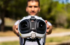 Una persona sostiene un casco polivalente blanco de la marca POC Amidal, con grandes aberturas de ventilación negras orientadas hacia la cámara, al aire libre y con vegetación al fondo, lo que lo hace ideal para el ciclismo de carretera.