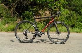 Una bicicleta de carretera negra y roja, con manillar de carretera y ruedas Zipp, está aparcada en la acera; lleva neumáticos Schwalbe Pro One Aero montados en la rueda delantera y la rueda trasera, con vegetación y arbustos al fondo.