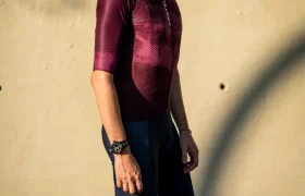 Un ciclista que lleva un casco blanco, gafas de sol, un maillot marrón rojizo Castelli Climber’s A/C y unos pantalones cortos tipo culotte azul marino A/C se encuentra de pie en una calle bañada por el sol, junto a una pared de color beige, preparado para dar el máximo rendimiento en condiciones de calor.