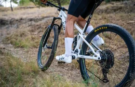 Un ciclista con pantalones cortos negros sube en una bicicleta de montaña blanca por un sendero de tierra cubierto de hierba. Para demostrar su rendimiento de gama alta, el ciclista lleva zapatillas y calcetines blancos Northwave Extreme X, que le aportan mayor versatilidad en terrenos exigentes.