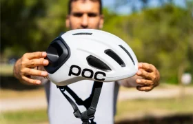 Una persona sostiene al aire libre un casco de ciclismo polivalente POC blanco con rejillas de ventilación negras y una correa para la barbilla, con árboles y césped al fondo; perfecto tanto para salidas por carretera como por grava.