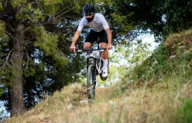 Una persona que lleva casco y gafas de sol recorre en bicicleta de montaña, equipada con un manillar integrado de carbono FSA NS SIC, un sendero de tierra rodeado de hierba y árboles.