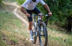 Un ciclista, que lleva casco y gafas de sol, pedalea en una bicicleta de montaña equipada con ruedas FSA cuesta arriba por un estrecho sendero de tierra rodeado de vegetación, demostrando un control total sobre el exigente terreno.