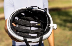 Una persona sostiene ante la cámara un casco de bicicleta blanco «Casco Polivalente», mostrando el acolchado interior y una etiqueta de seguridad amarilla con la inscripción «Mips» visible en el interior. El fondo es una zona al aire libre con césped, perfecta para aventuras en gravel o carretera.